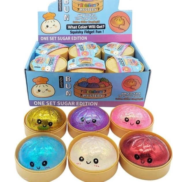 Squishy bolinho com glitter em cestinho, em várias cores, com caixa expositora Rainbow Mystery ao fundo (a caixa não acompanha o produto)